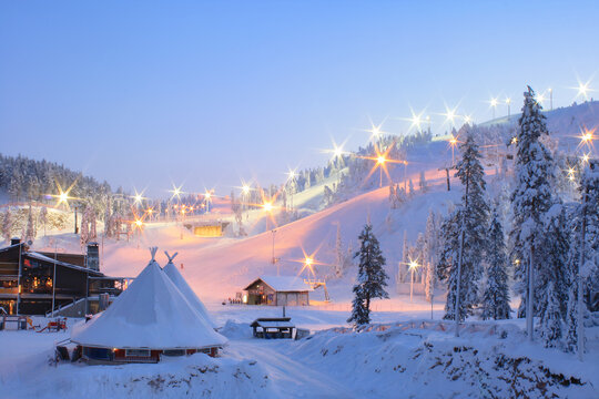 Picturesque winter sunrise at Ruka Ski Resort in Finland, with snow-covered slopes, glowing lights, frosted trees and magical morning atmosphere in a Nordic landscape.