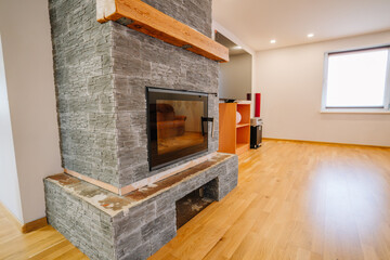 Modern living room featuring a stylish stone fireplace with wooden mantel, warm wooden flooring, and natural light streaming through a large window, creating a cozy atmosphere