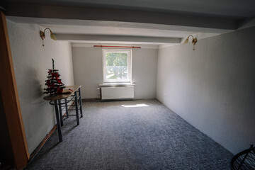 Empty room with gray walls and carpet, featuring a small Christmas tree on a table, bright natural light streaming through the window, creating a cozy atmosphere with copy space
