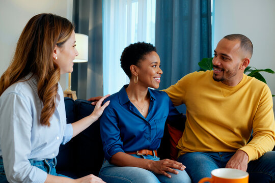 Young woman, young Black woman, young man sitting together on sofa smiling and talking, friendly interaction among diverse group of friends indoors