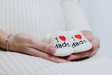 Hands of a pregnant Caucasian girl holding baby shoes with the inscription I love dad.