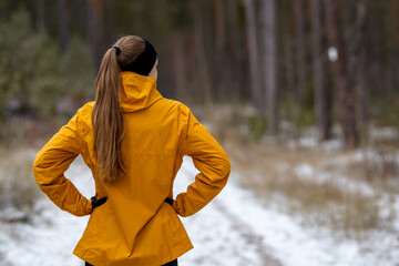 View from the back of a woman hiker pausing with hands on hips on a snowy path. Healthy lifestyle concept, enjoying a solitary winter walk or nature contemplation.