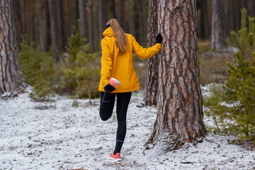 Sporty woman holding a pine tree while stretching in a snow-covered forest, concept of cold weather...