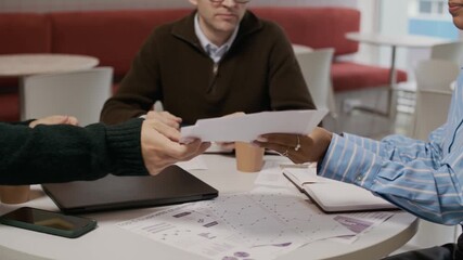Three colleagues sitting at table with coffee cups, laptop and documents on top. They looking at last report, trying to discuss it