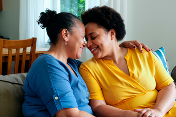 Adult Black women sitting close together on sofa smiling and embracing, touching foreheads and sharing affectionate moment in home setting, both appearing joyful and relaxed