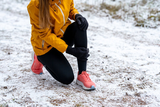 Female athlete forced to stop during a winter run, bending down and clutching her lower leg and knee. Image represents overuse injury, shin splints, and the risks of training in cold conditions.