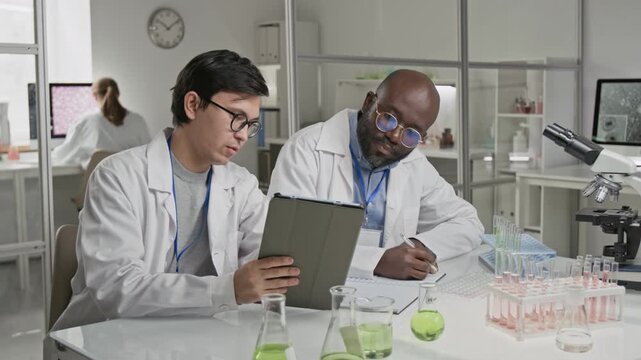 Two multiracial scientists in lab coats working with documents together, sitting at desk in laboratory