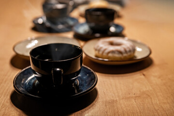 Several dark ceramic coffee cups and saucers with powdered sugar donut on a wooden table