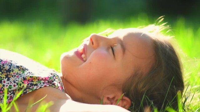Smiling happy little girl kid lying on green grass and dreaming. Cute six years old child enjoying nature outdoors. Healthy carefree kid playing outside in summer park on a lawn. Slow motion