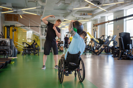 Man in wheelchair do exercise with weighted ball to tilt torso, lift with single arm press against neck. Trainer explain which muscles to engage to execute movement correctly, cervical resistance work