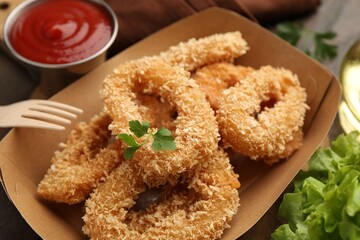 Deep fried squid rings with parsley in takeaway paper box, lettuce and ketchup on wooden table, closeup