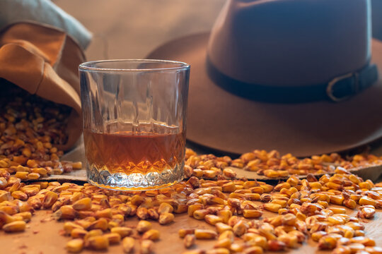 Low-angle, moody close-up of a glass of amber whiskey or bourbon on a wooden surface, surrounded by corn kernels, with a blurred cowboy hat and burlap sack in the background