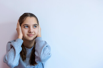 Teen girl covering ears with thoughtful expression. A young girl in a gray sweatshirt covers her ears while looking up, suggesting noise avoidance, deep thought, or sensory overload. Copy space.
