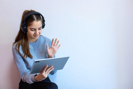 Teen girl video chatting on tablet with headphones. A smiling teenage girl wearing headphones waves at the tablet screen, engaged in a video call or online learning session from home.