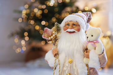Santa Claus figure with sack of gifts and toy bear against blurred lights indoors, closeup with...
