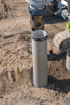 Overhead view of dusty construction site showing heavy machinery with large drilling rig and steel casing segments being installed for deep pile foundations in excavated earth.