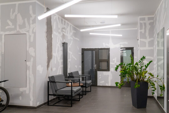 Modern minimalist waiting area with industrial-chic unfinished walls, sleek linear lighting, and comfortable seating, featuring vibrant green plant and dark framed doors.