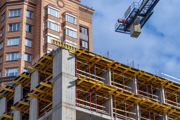 New multi-story concrete building under construction featuring intricate yellow formwork and scaffolding, next to finished brick residential tower, all beneath clear blue sky with prominent tower cran