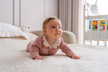 Sweet baby girl in charming pink knitted outfit with ruffled collar practicing tummy time on soft bed, gazing up in bright nursery