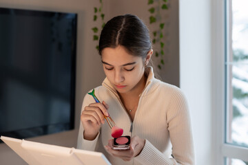 Teen girl applying makeup at home by window. A focused teenage girl uses a blush brush and mirror while doing her makeup in a cozy indoor setting with natural light.