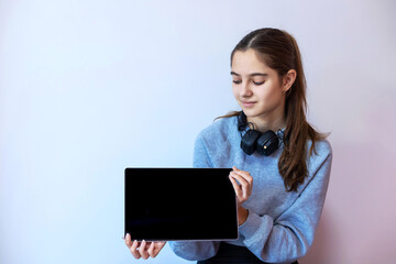 Teen girl showing blank tablet screen indoors. A smiling young girl with headphones around her neck presents a black tablet screen while sitting against a light-colored wall.