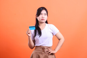 Naklejka premium A pensive woman holding a credit card while looking skeptically, suggesting uncertainty or concern about a financial transaction, isolated on orange background.