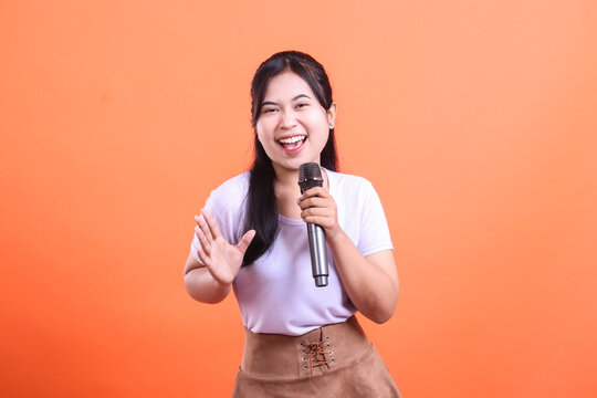 A cheerful young woman sings into a microphone with a big smile, her hand raised in a welcoming gesture. isolated on orange background