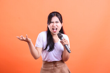 A young woman yells into a microphone with an angry expression, she is gesturing a question with her left hand, isolated on orange background
