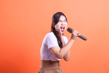 A young asian woman enthusiastically sings into a microphone, with her mouth open and hand near her face; isolated on orange background