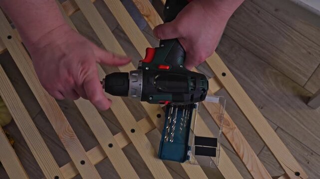 Hands selecting drill bit from accessory set case next to cordless drill on wooden shelf rack top view. Concept of choosing right tool size, DIY preparation and power equipment accessories usage.