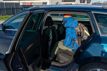 Boy with backpack getting into car back seat through open door in blue winter jacket on sunny day in suburban area. Concept of school commute, family transportation, daily routine and child safety.