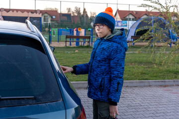 Boy in blue winter jacket and glasses standing near car looking back at playground in suburban area on autumn day. Concept of leaving after outdoor play, childhood moments and family transportation.