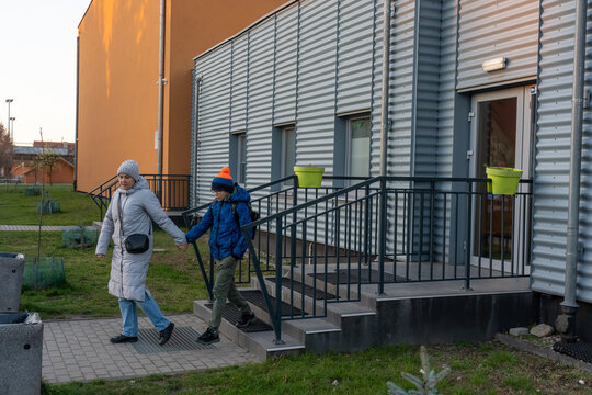Mother and son holding hands walking down stairs from modern building entrance in winter clothes at dusk. Concept of after school pickup, family bonding, parenting care and daily routine lifestyle. 