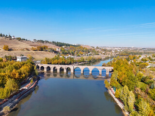 Fototapeta premium Stunning aerial view of the historic Ten-Eyed Bridge (On Gözlü Köprü) over the Tigris River. The stone arches and their reflection are framed by vibrant autumn trees under a bright blue sky.