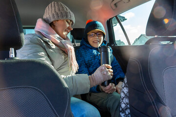 Happy mother and son sitting in car back seat holding thermos with hot drink on cold winter day. Concept of family road trip, winter travel, togetherness and cozy moments during automobile journey. 