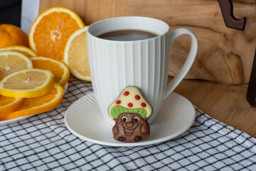 Cute chocolate mushroom character positioned on white saucer next to coffee cup with fresh orange and lemon slices on wooden cutting board. Concept of cheerful breakfast and citrus coffee pairing. 
