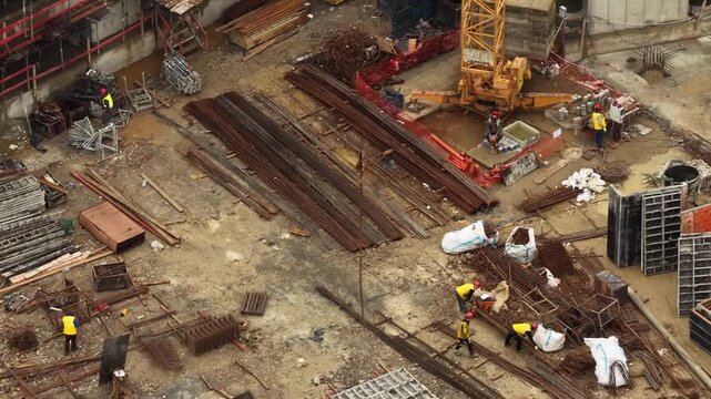 Aerial orbital shot around a crane hook chain with construction workers organizing rebar and formwork materials on a busy building site