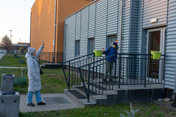 Mother waving goodbye to son with backpack standing on stairs of modern building entrance at dusk in winter clothes. Concept of school drop-off, family bonding, parenting routine and childhood care.