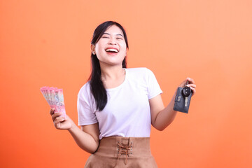 A cheerful woman is holding a car key and a fan of money, expressing excitement and satisfaction with her new purchase isolated on orange background.