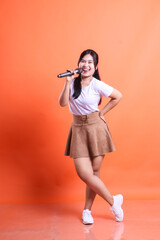 A beautiful Asian woman enjoys singing with a microphone in a studio, exuding happiness and joy. isolated on orange background.