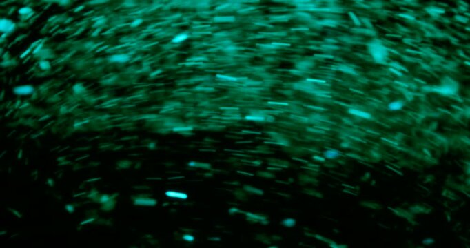 Particles float in water over a dark background, reflecting top light in green and blue hues, creating a glittering, halo-like, cosmic universe effect.