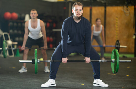 Focused motivated young bearded man performing barbell deadlift to increase strength and stability of back, glutes and hamstrings during intense cross training at gym