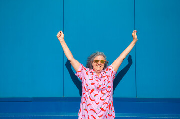 Happy senior woman raising arms celebrating success in front of blue wall