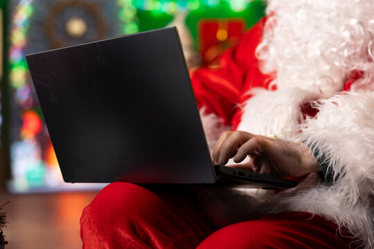 Close up of Santa Claus using laptop in festive log cabin to navigate online webpages. Father Christmas in North Pole workshop browsing internet on notebook, checking holiday gift lists