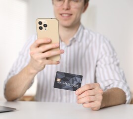 Young man making online payment with phone and credit card at white table indoors, closeup