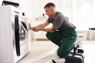 Smiling repairman in uniform fixing broken washing machine with screwdriver at home