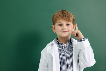 Cute little boy in laboratory coat pointing upwards near green chalkboard, space for text. Child...