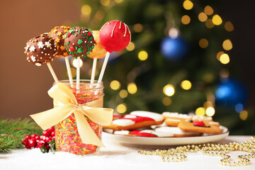 Sweet cake pops with sprinkles in jar, gingerbread cookies and Christmas decor on table against blurred lights, closeup. Space for text