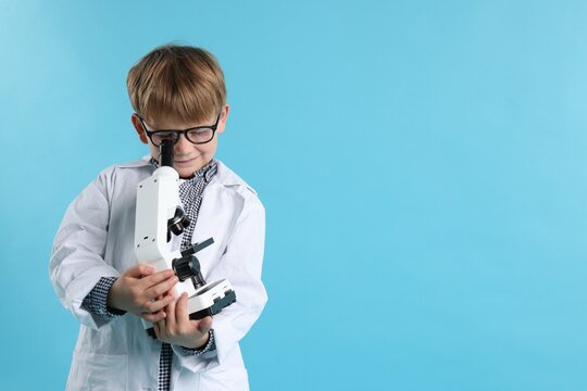 Cute little boy in laboratory coat and glasses with microscope on light blue background, space for text. Child and science