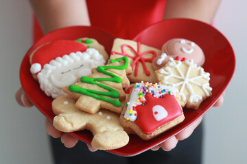 Hands holding a heart-shaped red plate filled with beautifully decorated shortbread and gingerbread cookies for Christmas and the holiday season.